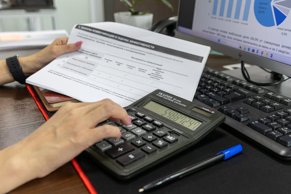 A person calculates financial data using a calculator and document, working at an office desk.